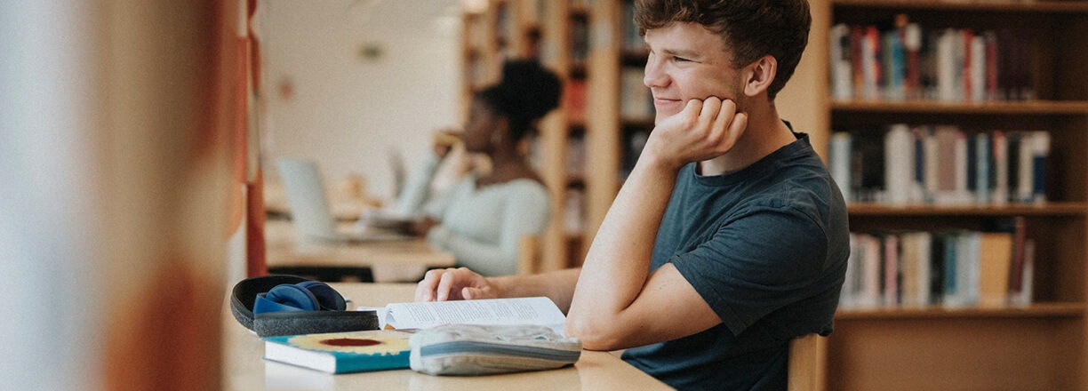 Ein Student, der an einem Schreibtisch mit Büchern und einem Laptop in einer Bibliothek lernt.