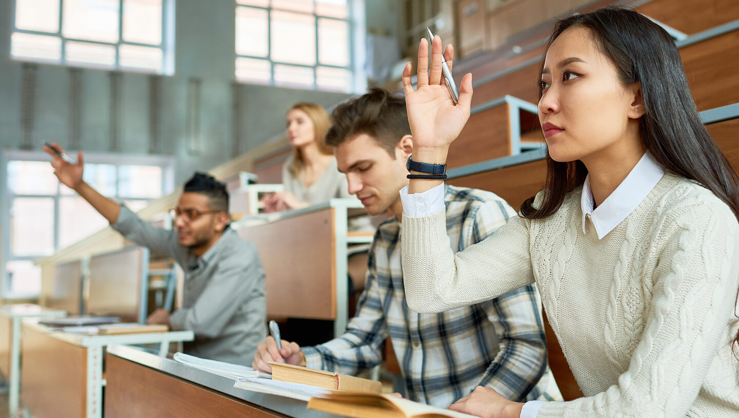 Ein Universitätsraum mit Studierenden, die an Schreibtischen sitzen, während eine Studentin im Vordergrund die Hand hebt.